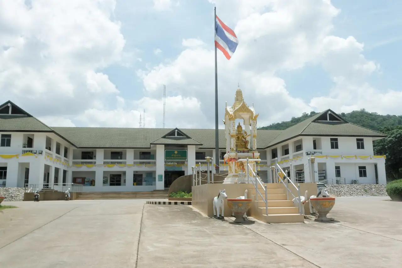 อาคารที่ว่าการเทศบาลปากช่องที่มีศาลพระพรหมและเสาธงหน้าอาคาร ชื่อไฟล์บนคอมมอนส์ระบุว่าเป็นสำนักงานเทศบาลปากช่อง ภาพประกอบตัวเมืองปากช่องที่ใช้เป็นฐานก่อนไปฟาร์ม ไม่ใช่ภาพในฟาร์ม ภาพจาก Wikimedia Commons ผู้ถ่าย Ahoerstemeier สัญญา CC BY 4.0