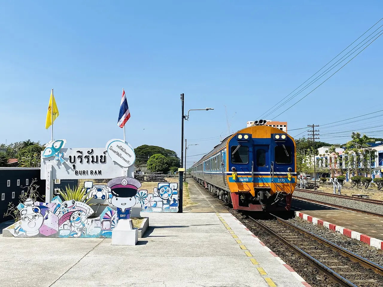 ขบวนรถดีเซลรางปรับอากาศที่สถานีรถไฟบุรีรัมย์ มีป้ายชื่อสถานีและตุ๊กตาการ์ตูนเจ้าหน้าที่การรถไฟ ภาพจาก Wikimedia Commons ผู้ถ่าย Reversemos.sapanaht ใช้สัญญา CC0 ชื่อไฟล์ระบุเป็นขบวนด่วนพิเศษที่ 21 กรุงเทพฯ ไปอุบลราชธานี ภาพนี้เป็นภาพประกอบ ไม่ใช่ตารางเดินรถปัจจุบัน