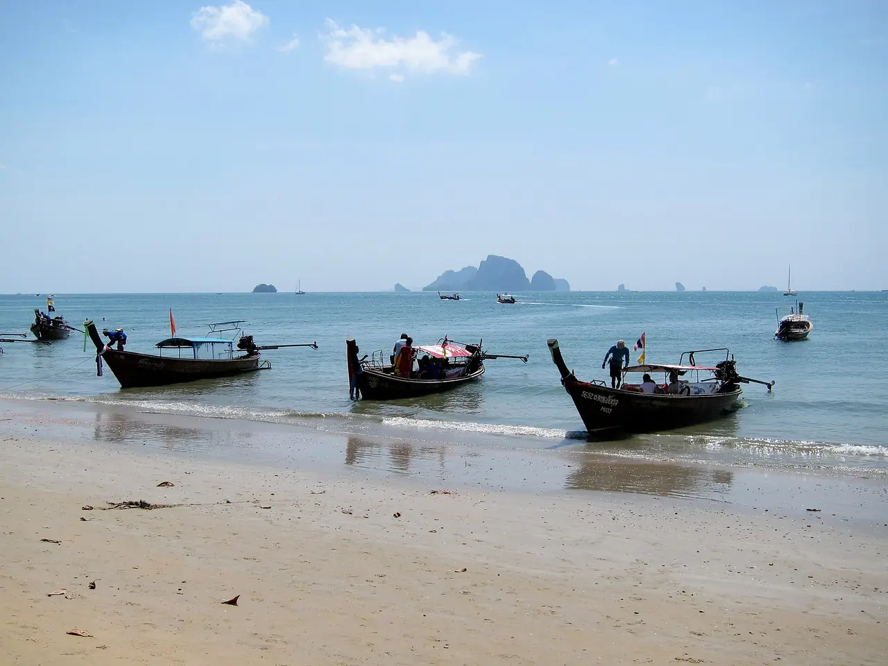 เรือหางยาวจอดที่ชายหาดอ่าวนางพร้อมเกาะหินปูนเล็ก ๆ ไกลออกไปกลางทะเล ภาพจาก Wikimedia Commons ผู้ถ่าย Jukka from HELSINKI, Finland สัญญา CC BY 2.0 ไม่ระบุปีที่ถ่าย เป็นภาพประกอบ เรือและป้ายอาจเปลี่ยนไปแล้ว