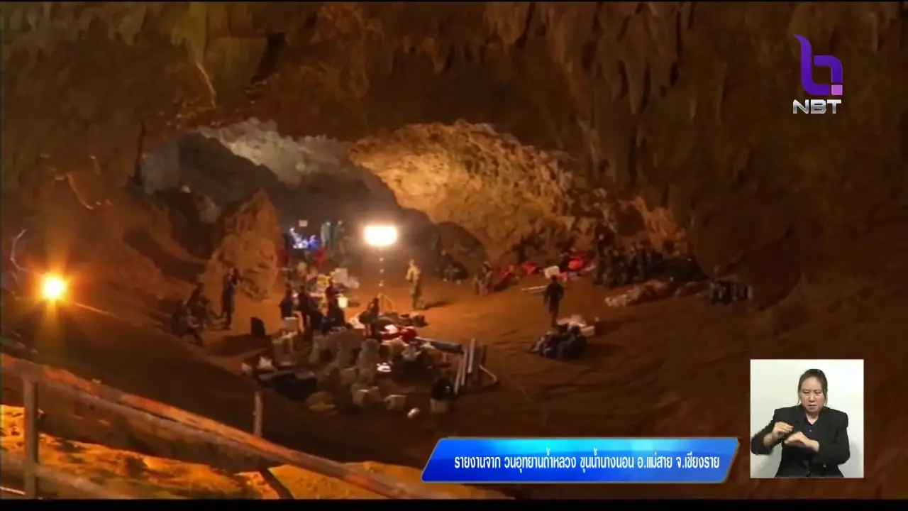 ภาพนิ่งจากรายการข่าวของ NBT แสดงกลุ่มคนและอุปกรณ์ในโถงทางเข้าถ้ำหลวง แถบข่าวด้านล่างลงชื่อวนอุทยานถ้ำหลวง ขุนน้ำนางนอน อ.แม่สาย จ.เชียงราย ภาพจาก Wikimedia Commons ผู้ถ่าย NBT สัญญา CC BY 3.0 ชื่อไฟล์เกี่ยวกับปฏิบัติการปี 2561 แต่ไม่ลงวันที่ถ่ายในรายละเอียดที่เราอ่าน
