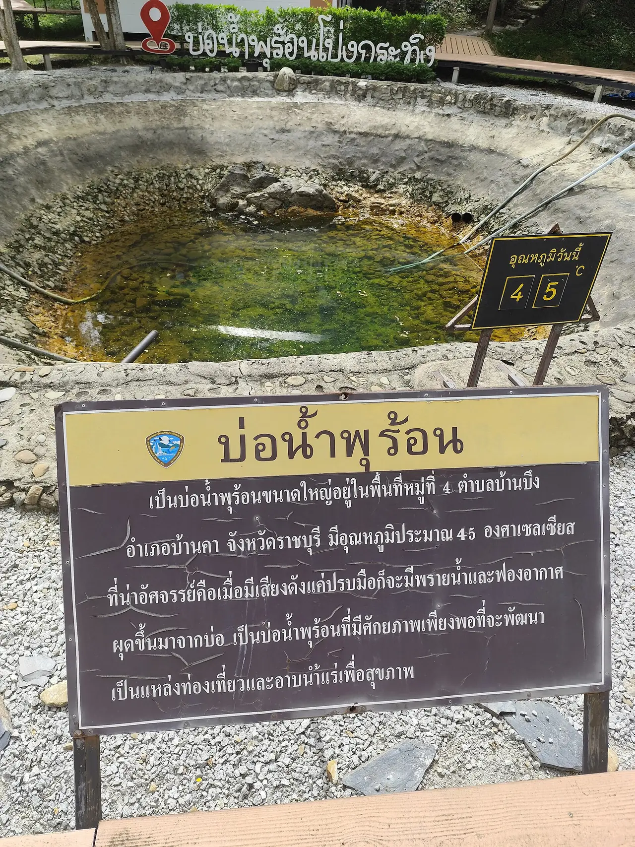 บ่อน้ำพุร้อนวงกลมขอบหินปูนมีสีเขียวเหลืองในน้ำ มีป้ายเขียนว่าบ่อน้ำพุร้อนโป่งกระทิง ป้ายวัดอุณหภูมิตอนถ่ายแสดงตัวเลข 45 และป้ายอธิบายบอกว่าบ่อนี้อยู่ที่ตำบลบ้านบึง อำเภอบ้านคา จังหวัดราชบุรี อุณหภูมิประมาณ 45 องศาเซลเซียส ไม่ใช่บ่อน้ำพุร้อนพระร่วง ใช้ให้เห็นรูปแบบบ่อน้ำพุร้อนท้องถิ่นที่มีป้ายอุณหภูมิ ภาพจาก Wikimedia Commons ผู้ถ่าย UNI279 สัญญา CC BY-SA 4.0 ถ่ายวันที่ 12 สิงหาคม 2568