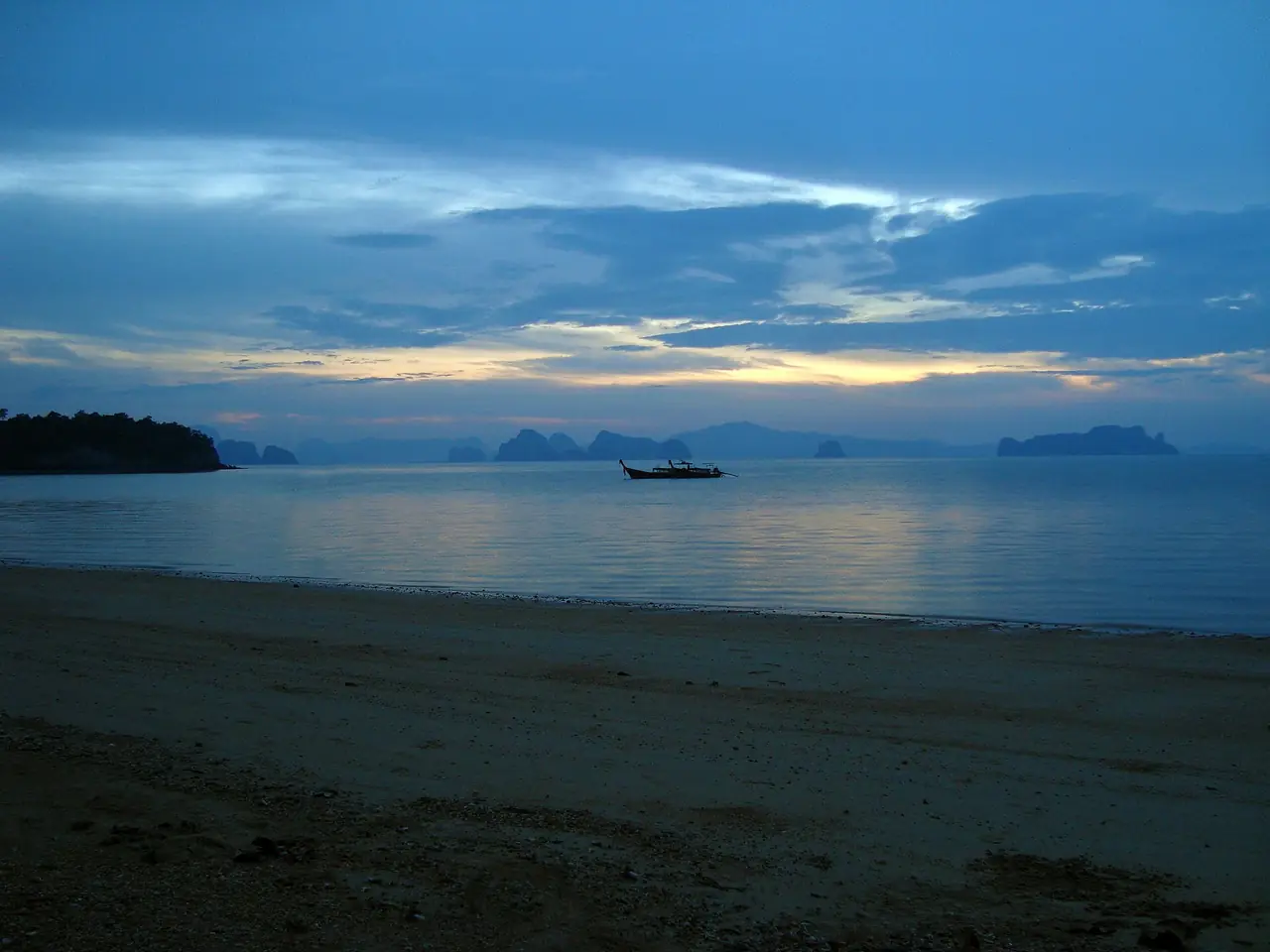 เรือหางยาวลำเดียวลอยในน้ำนิ่งตอนฟ้าสาง มองจากหาดบนเกาะยาวน้อยไปยังเกาะหินปูนไกล ๆ ภาพของ A J Butler จาก Wikimedia Commons สัญญา CC BY 3.0 ตามคำบรรยายภาพบน Commons เป็นพระอาทิตย์ขึ้นบนเกาะยาวน้อย ไม่ได้ระบุหาด ภาพเก่า