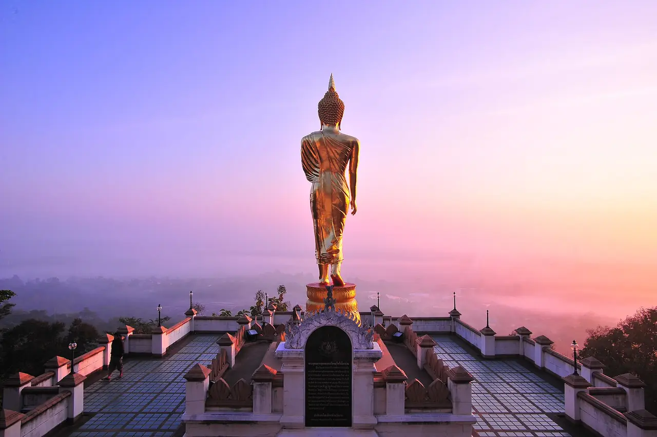 ลานชมวิวและพระพุทธรูปสีทองที่ฟ้าสีม่วงชมพูตอนรุ่งหรือใกล้ค่ำ ฐานพระมีซุ้มโค้งลายประดับ ลานปูกระเบื้องและมีคนยืนอยู่ด้านซ้าย ภาพจาก Wikimedia Commons ผู้ถ่าย K.Phothiwijit สัญญา CC BY-SA 3.0 ภาพนี้ไม่ลงเวลาถ่าย เราไม่ระบุว่าเป็นเช้าหรือเย็น เป็นภาพประกอบ