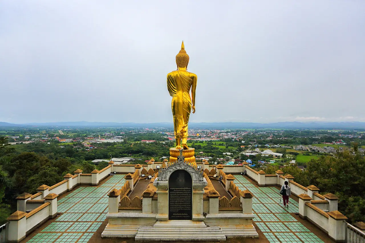 พระพุทธรูปสีทองมองจากด้านหลังบนลานกระเบื้องเขียวขาวตอนฟ้าครึ้ม มีคนยืนอยู่ที่ขอบลาน ด้านล่างเป็นทุ่งและหลังคาบ้านของตัวเมือง ภาพจาก Wikimedia Commons ผู้ถ่าย Yakuzakorat สัญญา CC BY-SA 4.0 ไม่ลงวันที่ถ่าย เป็นภาพประกอบ