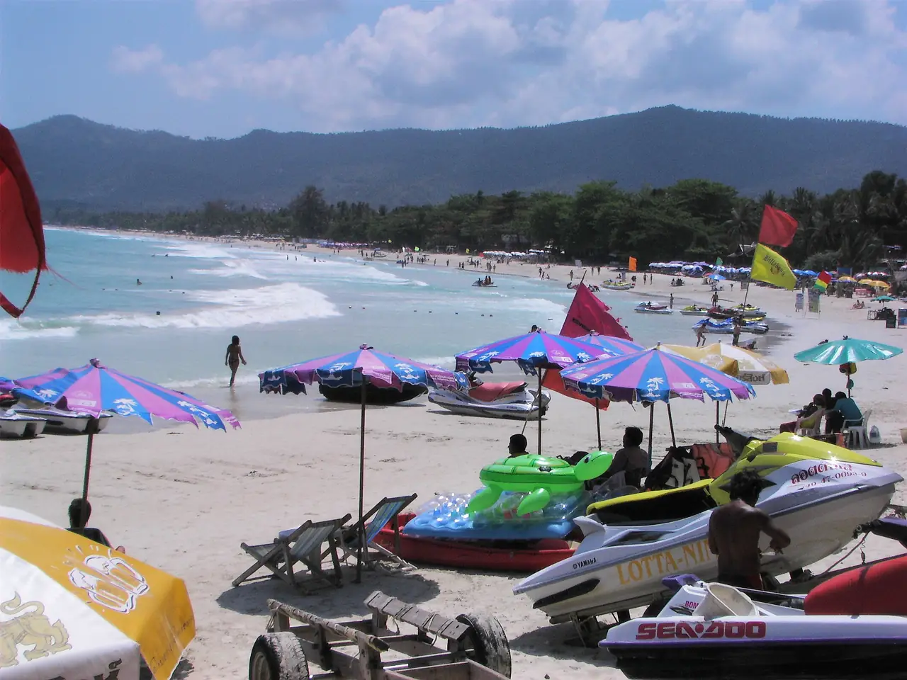 หาดเฉวงที่มีร่ม เจ็ตสกี และธงบนหาด ภูเขาอยู่เบื้องหลัง ภาพที่ Commons ระบุเป็นภาพสาธารณสมบัติจากผู้ใช้ Wipkinger ที่ Wikivoyage ไม่ทราบปีที่ถ่าย เป็นภาพเก่าใช้ประกอบ ไม่ใช่สภาพวันนี้ ภาพจาก Wikimedia Commons สัญญา สาธารณสมบัติ