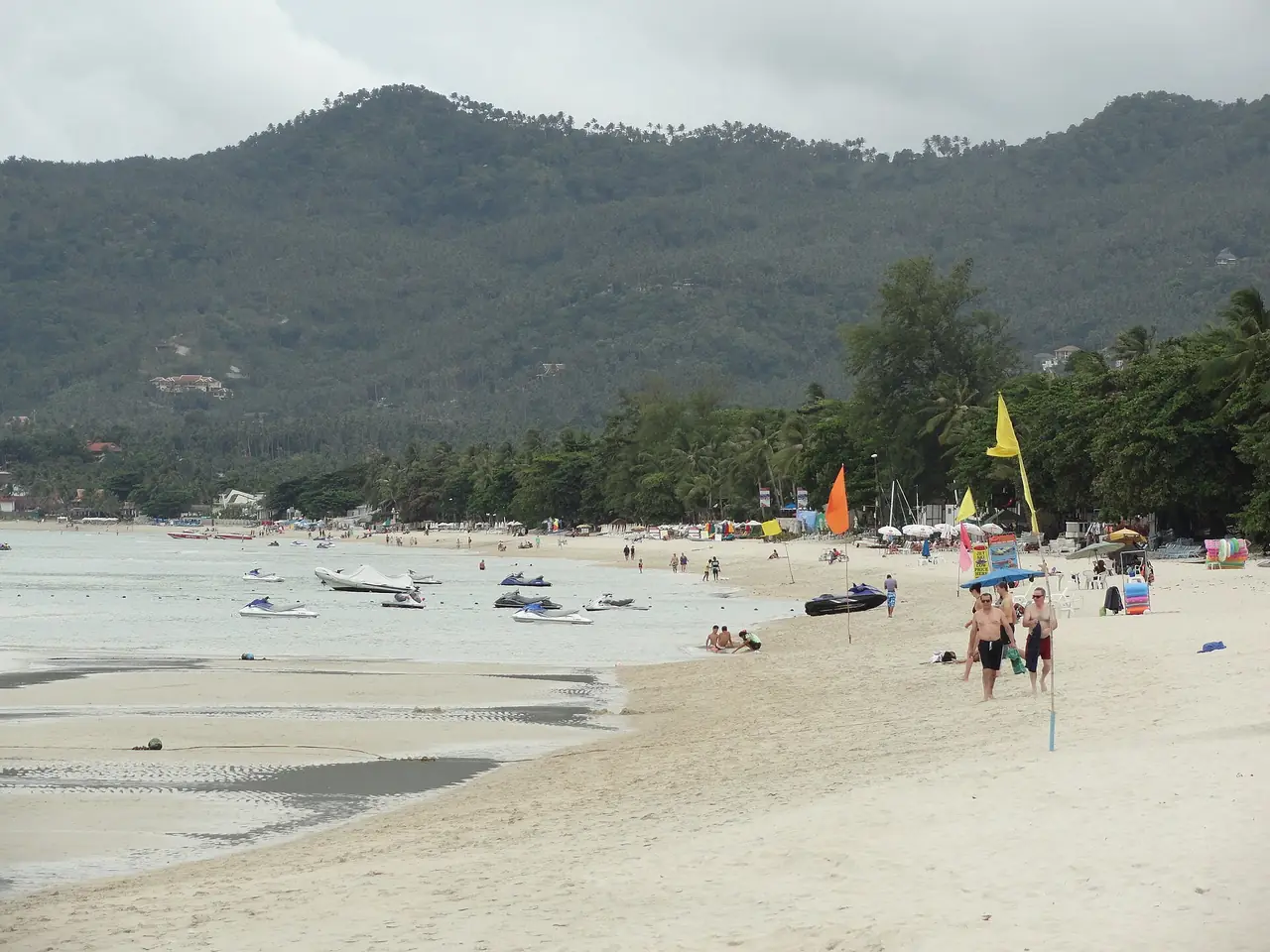 หาดเฉวงฝั่งที่มีเกลียวคลื่นเบา ฟ้าครึ้ม มีเรือเจ็ตสกีจอดบนหาดและภูเขาเขียวอยู่ด้านหลัง ภาพที่ Commons ติดป้ายหาดเฉวง ภาพนี้ถ่ายวันฟ้าครึ้ม แสดงสภาพวันเมฆมาก ไม่ใช่ฤดูใด ภาพจาก Wikimedia Commons ผู้ถ่าย Fabio Achilli สัญญา CC BY 2.0