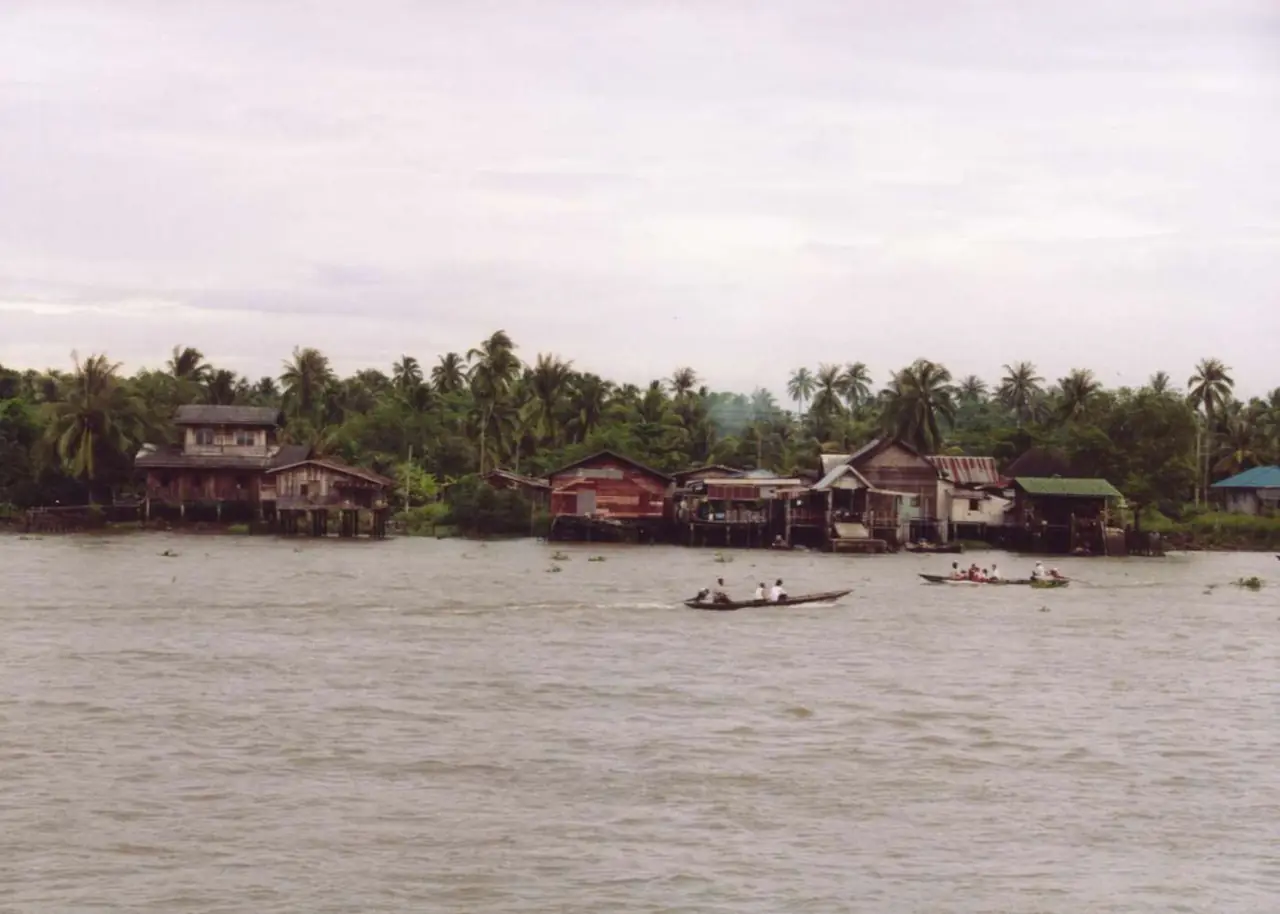 แม่น้ำตาปีที่สุราษฎร์ธานี เห็นบ้านเรือนริมน้ำใต้ต้นมะพร้าวและเรือหางยาวสองลำ ภาพจาก Wikimedia Commons ผู้ถ่าย Ahoerstemeier สัญญา CC BY-SA 3.0 ไม่ระบุปีที่ถ่าย ภาพออกโทนฟิล์มเก่า สภาพริมน้ำตอนนี้อาจต่างไป